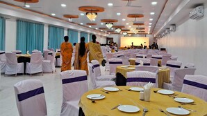 A banquet hall with round tables, white chairs, and a stage at the front. - Hotel Aaditya Inn Tathawade (Paud)