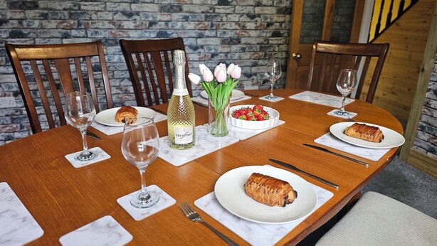Una mesa de comedor de madera dispuesta con platos, vasos, cubiertos, una botella de Freixenet, un florero con flores y un bol de fresas.