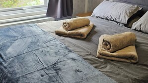 Un dormitorio con un ventanal grande, una cama con colcha gris, dos toallas enrolladas y un cabecero gris.