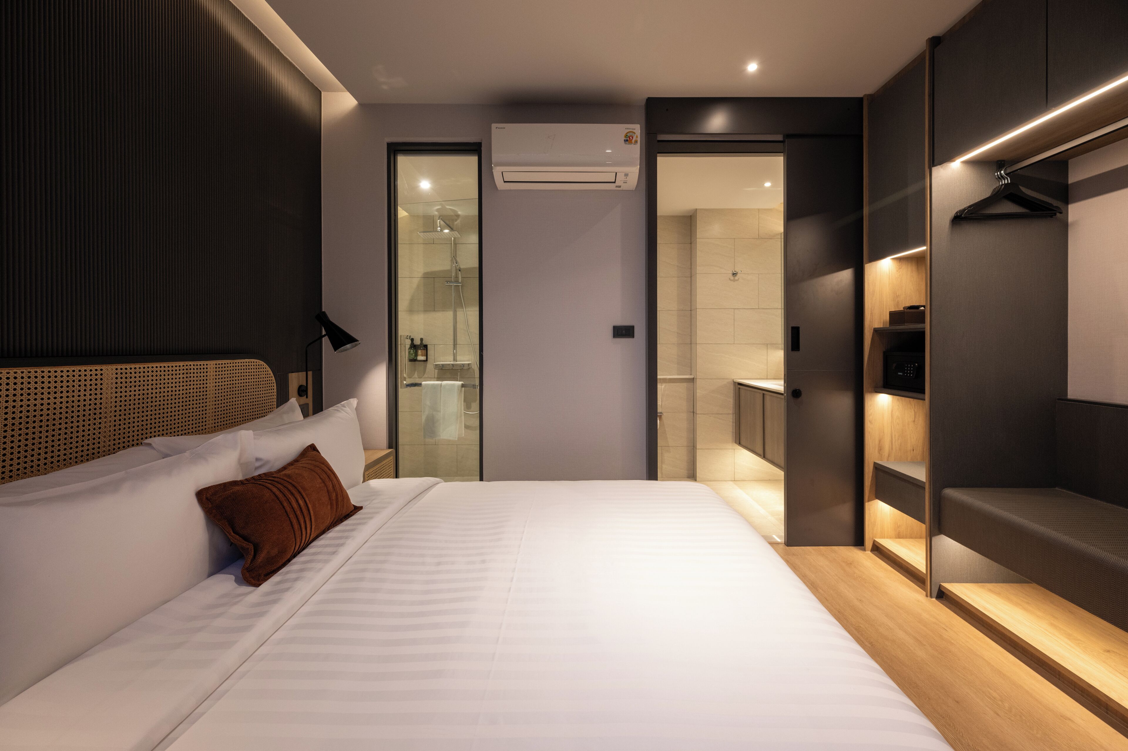 Una habitación de hotel moderna con una cama grande, un vestidor, un baño con ducha de vidrio y un equipo de aire acondicionado.