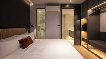 Una habitación de hotel moderna con una cama grande, un vestidor, un baño con ducha de vidrio y un equipo de aire acondicionado.