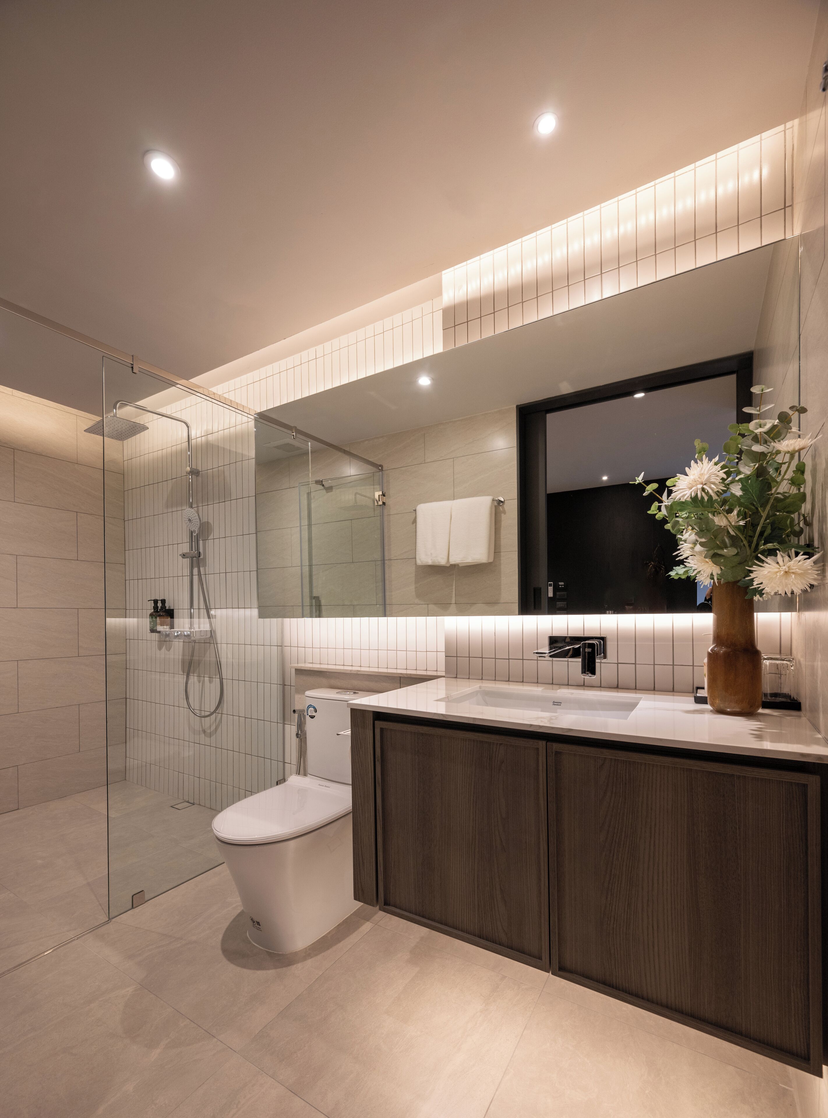 A modern bathroom with a glass shower enclosure, a toilet, a sink with a mirror above, and a vase with flowers on the countertop.