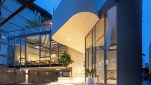A modern building with large glass windows, a curved entrance, and a well-lit outdoor area with steps leading to the entrance. - The Moment (Bangkok)