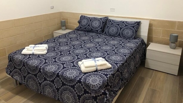 A neatly made bed with a patterned bedspread, two pillows, and two folded towels on the bed. There are two bedside tables with lamps. - Campomarino di Maruggio - Apartment in residence facing the sea - Monaco Mirante (Campomarino di Maruggio)