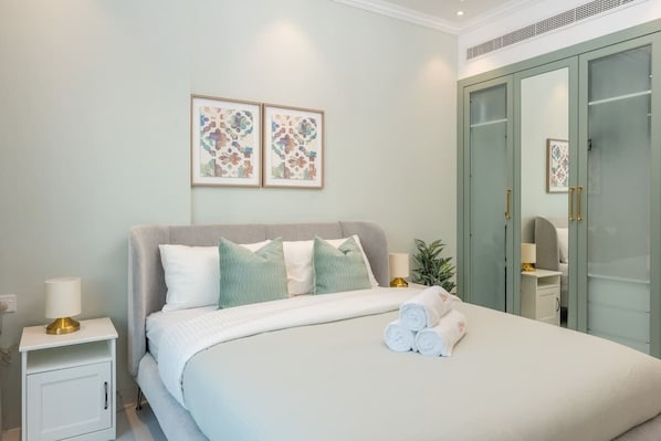 A neatly made bed with pillows, a bedside lamp, and rolled towels. There are two framed artworks on the wall and a wardrobe with a mirror.