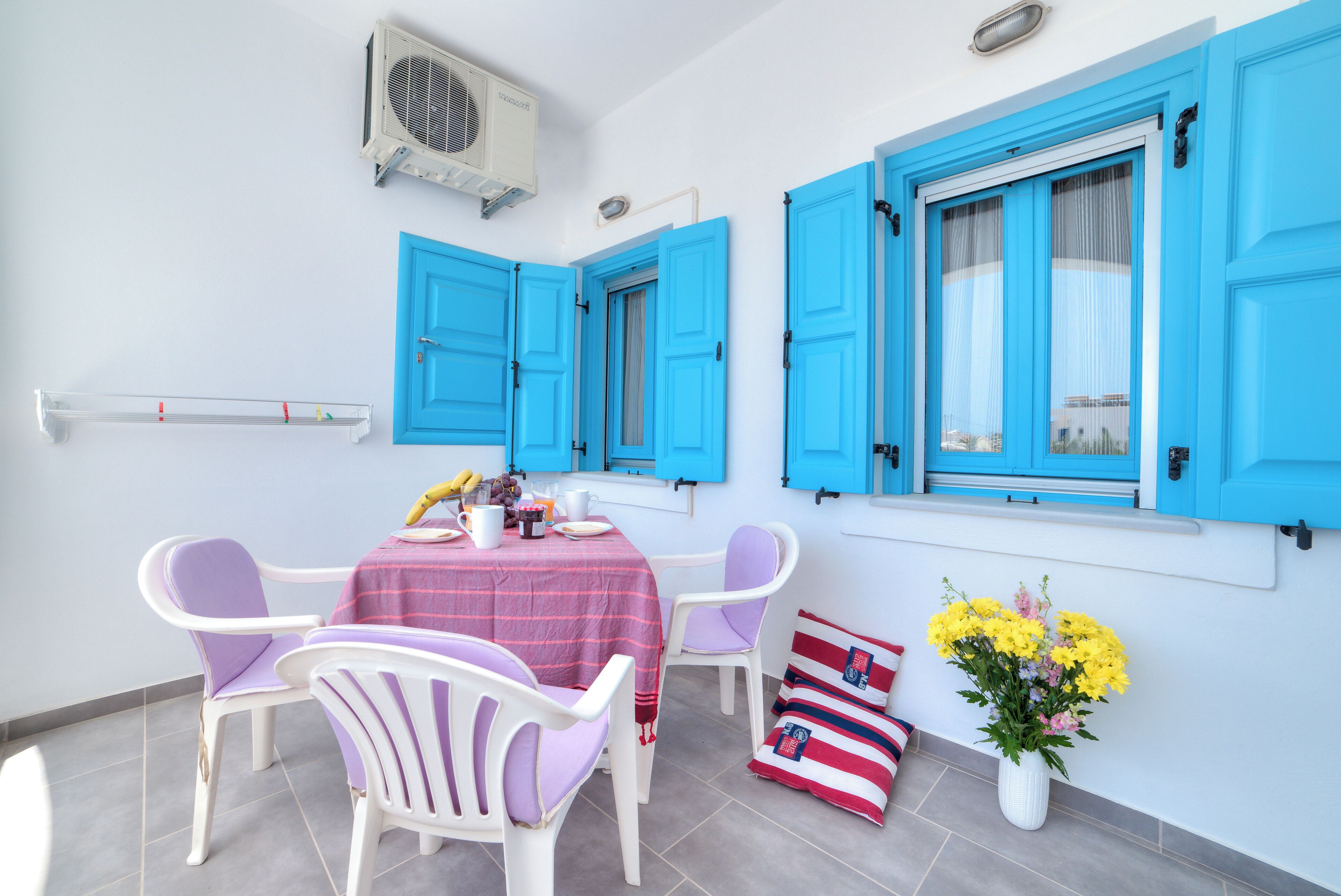A room with a table set for two, blue shutters, a white air conditioning unit, and a vase of yellow flowers.