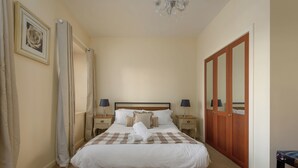 Ein ordentlich bezogenes Bett mit kariertem Überwurf und einem zusammengerollten Handtuch, zwei Nachttischlampen, ein Holzschrank und ein gerahmtes Bild an der Wand.