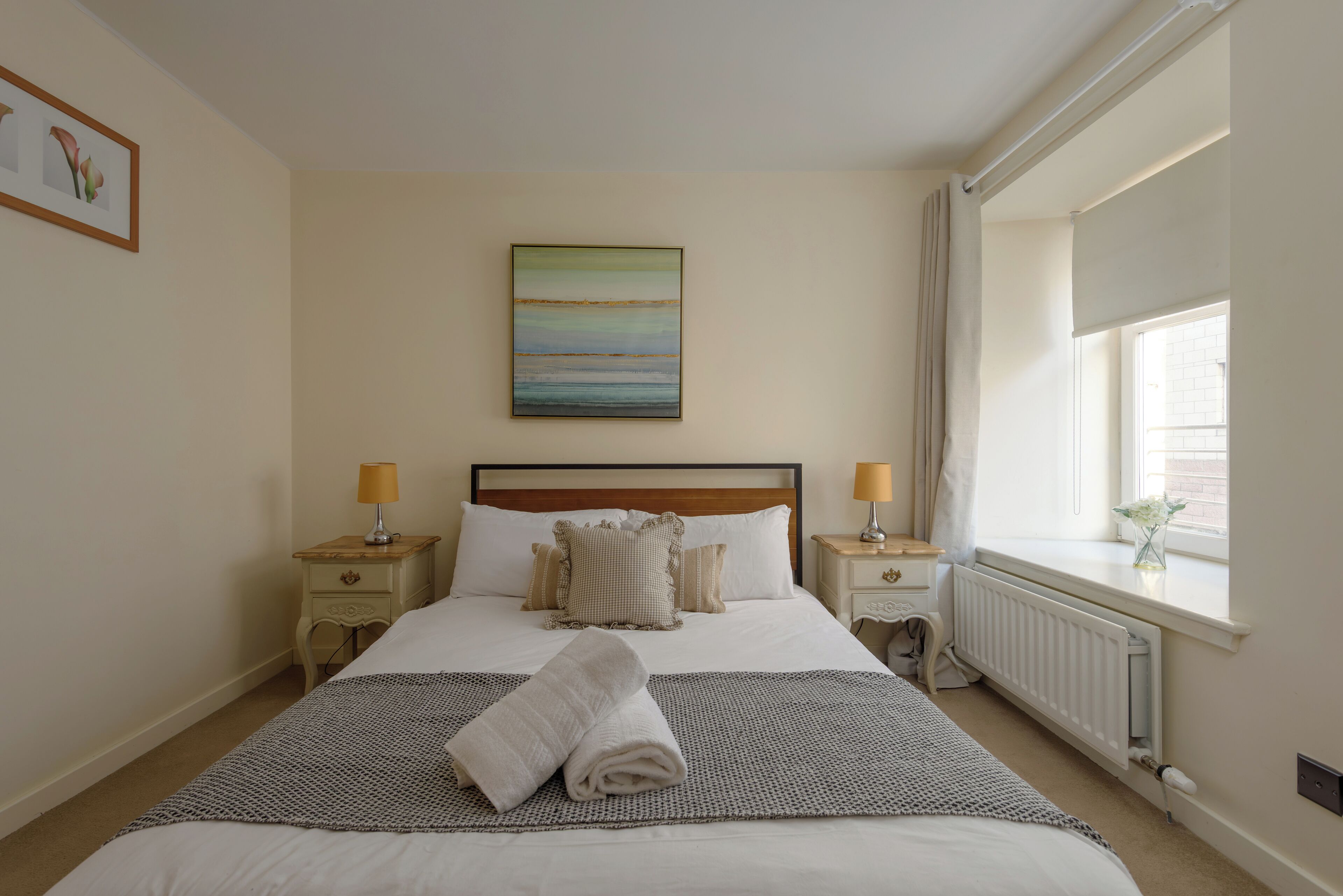 A neatly made bed with a folded towel, a painting above it, and a window with a view.