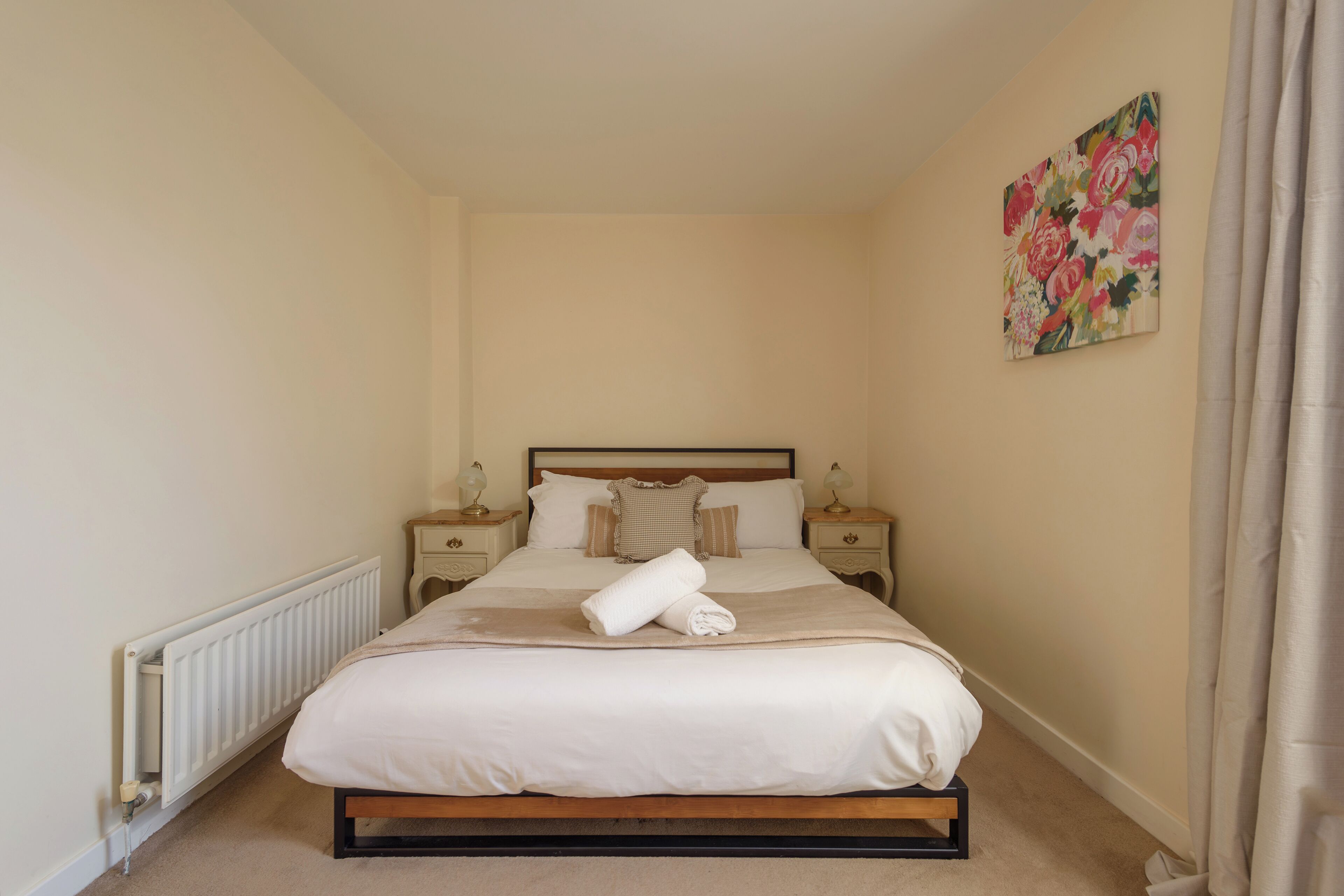 Un lit bien fait, avec une couette blanche et une serviette enroulée, une tête de lit en bois et un tableau floral accroché au mur.