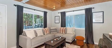 Una sala de estar acogedora con techo de madera, un sofá gris con cojines estampados, una mesa de centro y un otomano naranja pequeño.