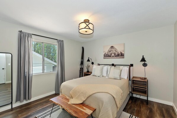 Um quarto com cama, mesa de cabeceira, luminária, janela com cortinas e um quadro na parede.