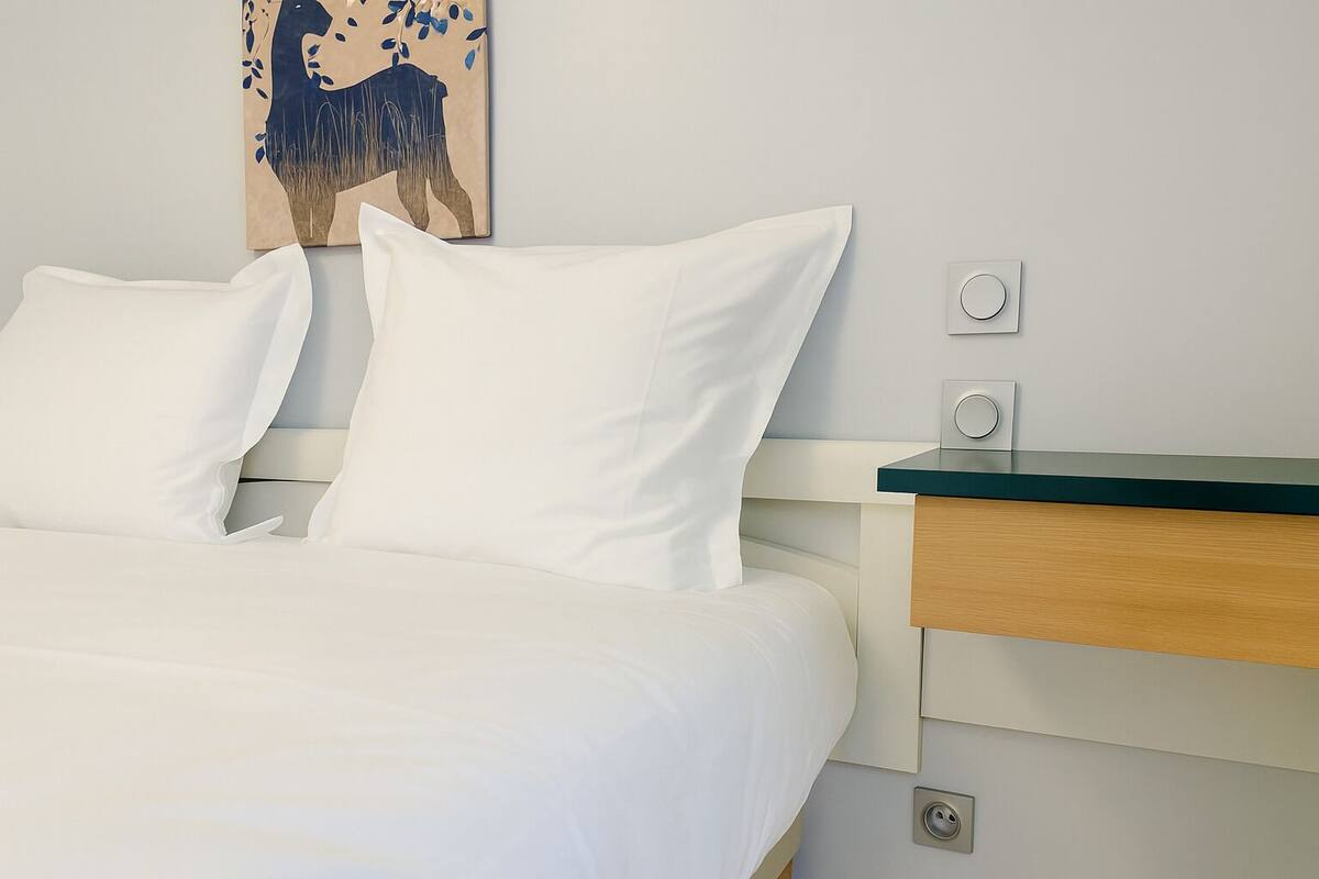 Un lit bien fait, avec du linge de lit blanc, une table de chevet en bois et un interrupteur fixé au mur.