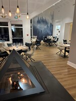 Un comedor moderno con mesas y sillas redondas, una chimenea y un mural de un bosque en la pared.