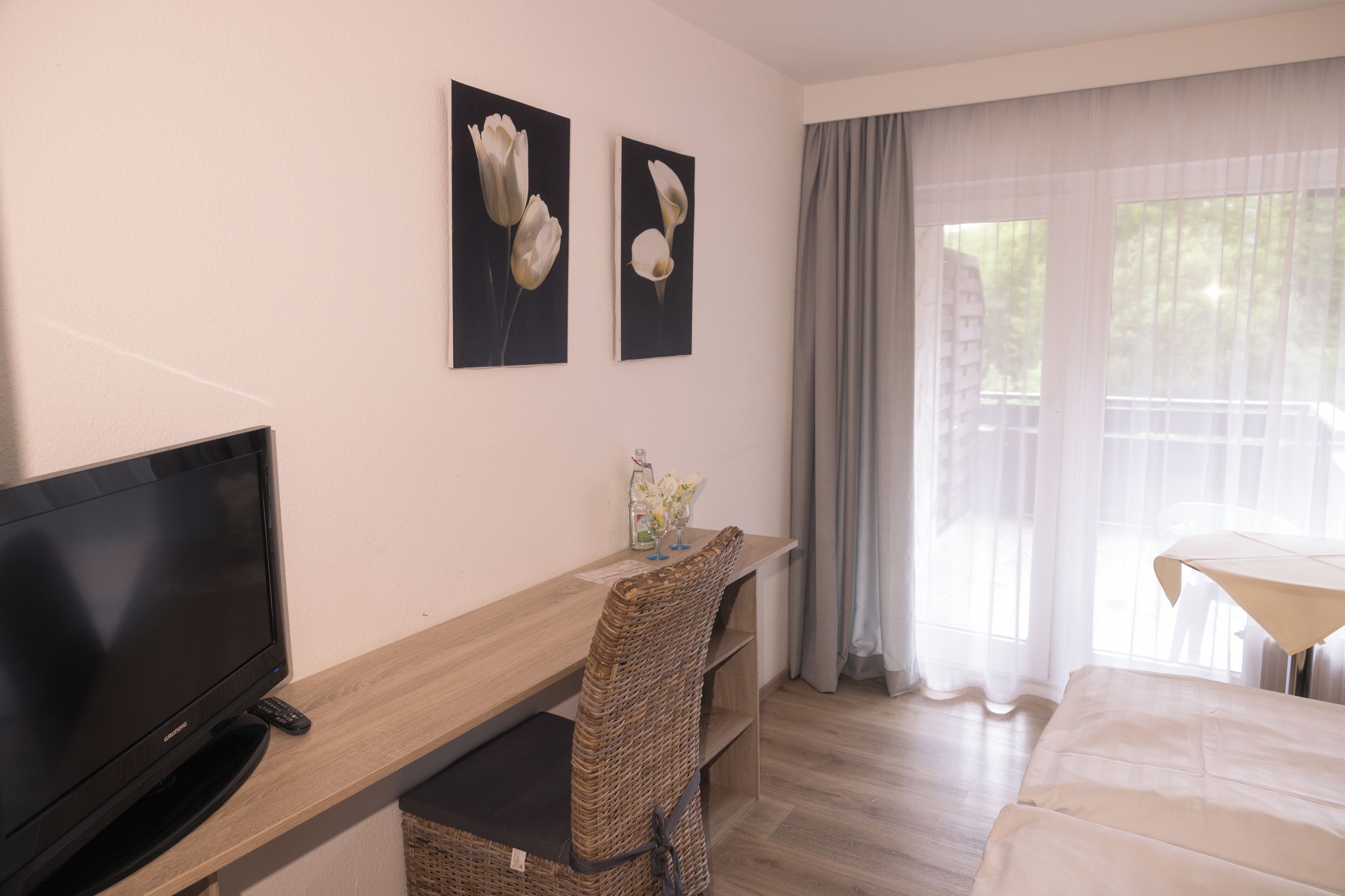 Habitación de hotel con televisión sobre un escritorio de madera, una silla de mimbre, una cama y dos cuadros de flores en la pared.