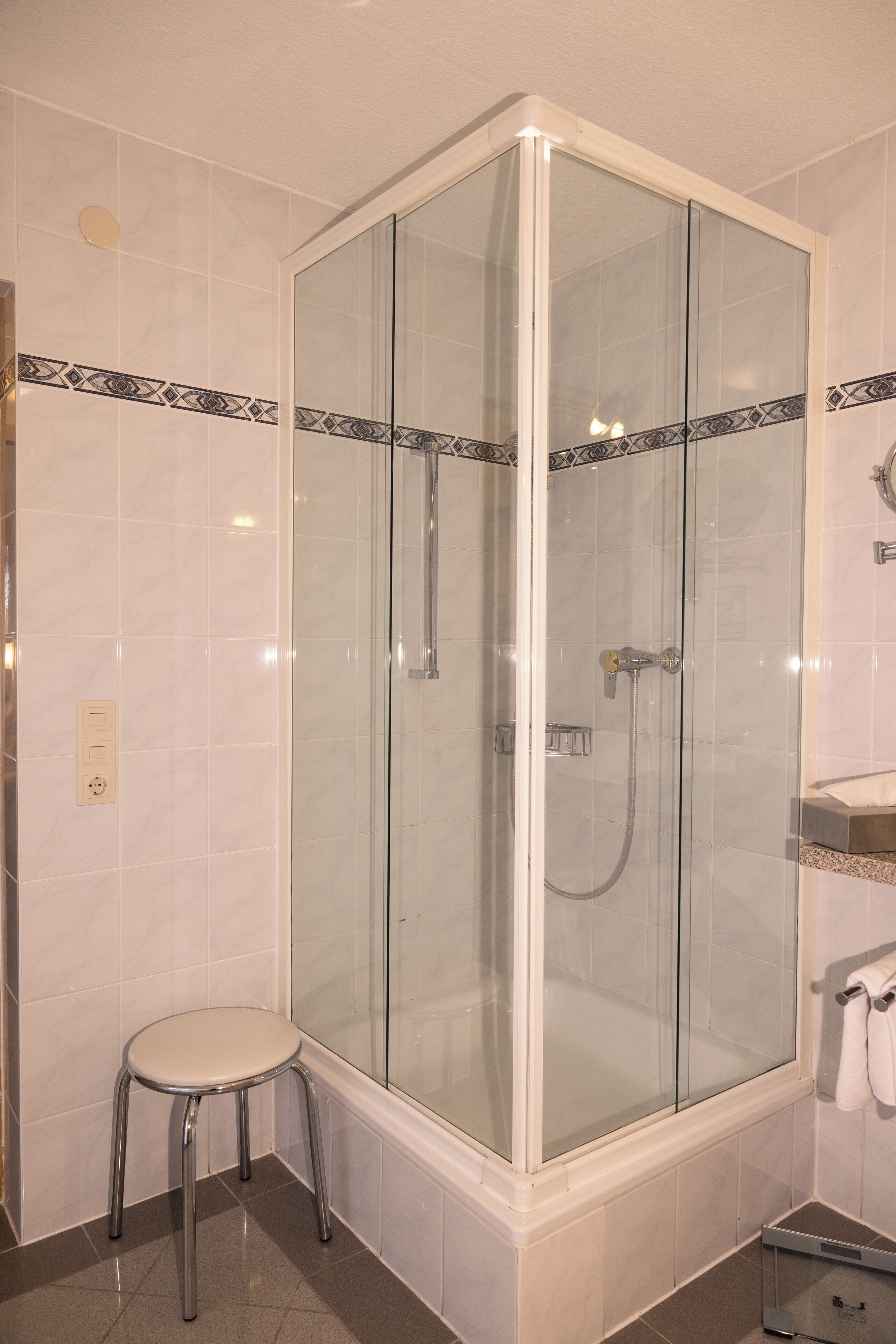 A glass-enclosed shower with a white tiled wall, a small stool, and a towel rack.