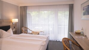 Ein Hotelzimmer mit einem Bett, einem Schreibtisch mit Lampe, einem Sessel, einem Tisch und einem Fenster mit durchsichtigen Vorhängen.