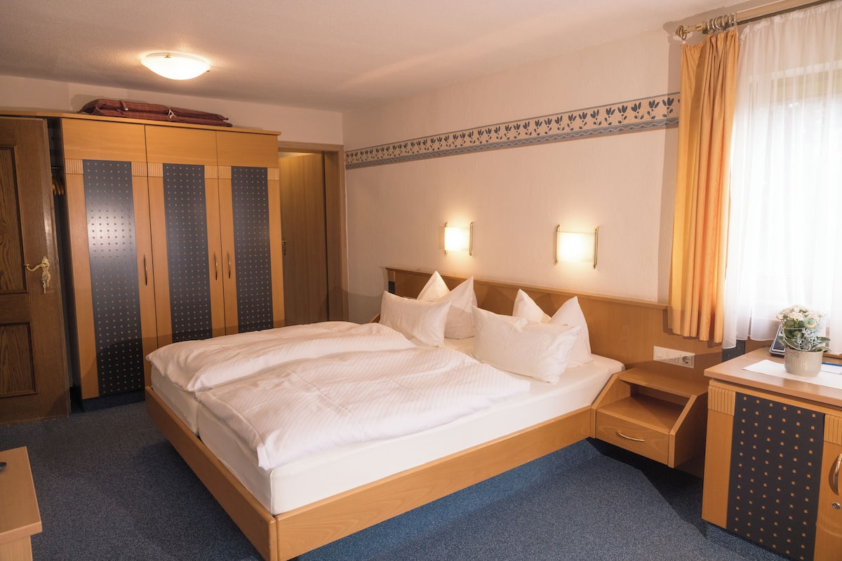 Habitació d'hotel amb un llit gran, un armari de fusta, taules de nit i un petit escriptori amb una planta.