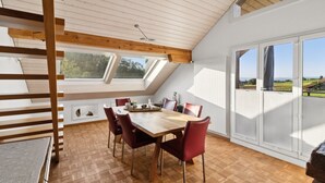 A dining area with a wooden table and red chairs, large windows with a view of the countryside, and a sloped ceiling with wooden beams.