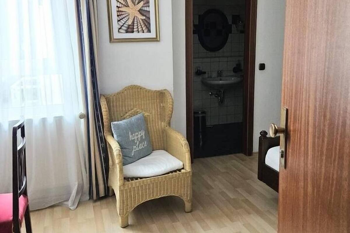 A room with a wicker chair, a bed, a bathroom with a sink, and a door leading to another room.