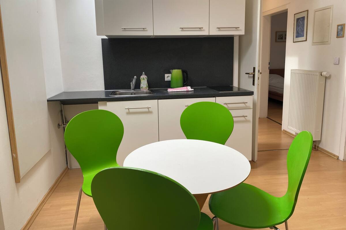 A compact kitchen with white cabinets, a black countertop, and a small dining table with green chairs.