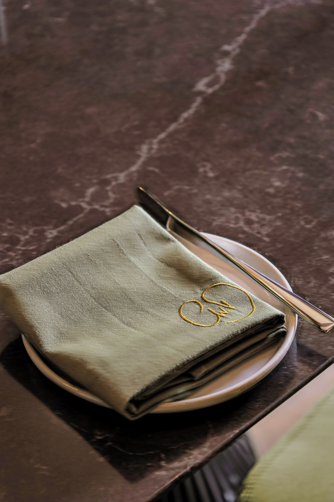 A folded green cloth napkin with a gold embroidered design on a plate, accompanied by a silver fork.