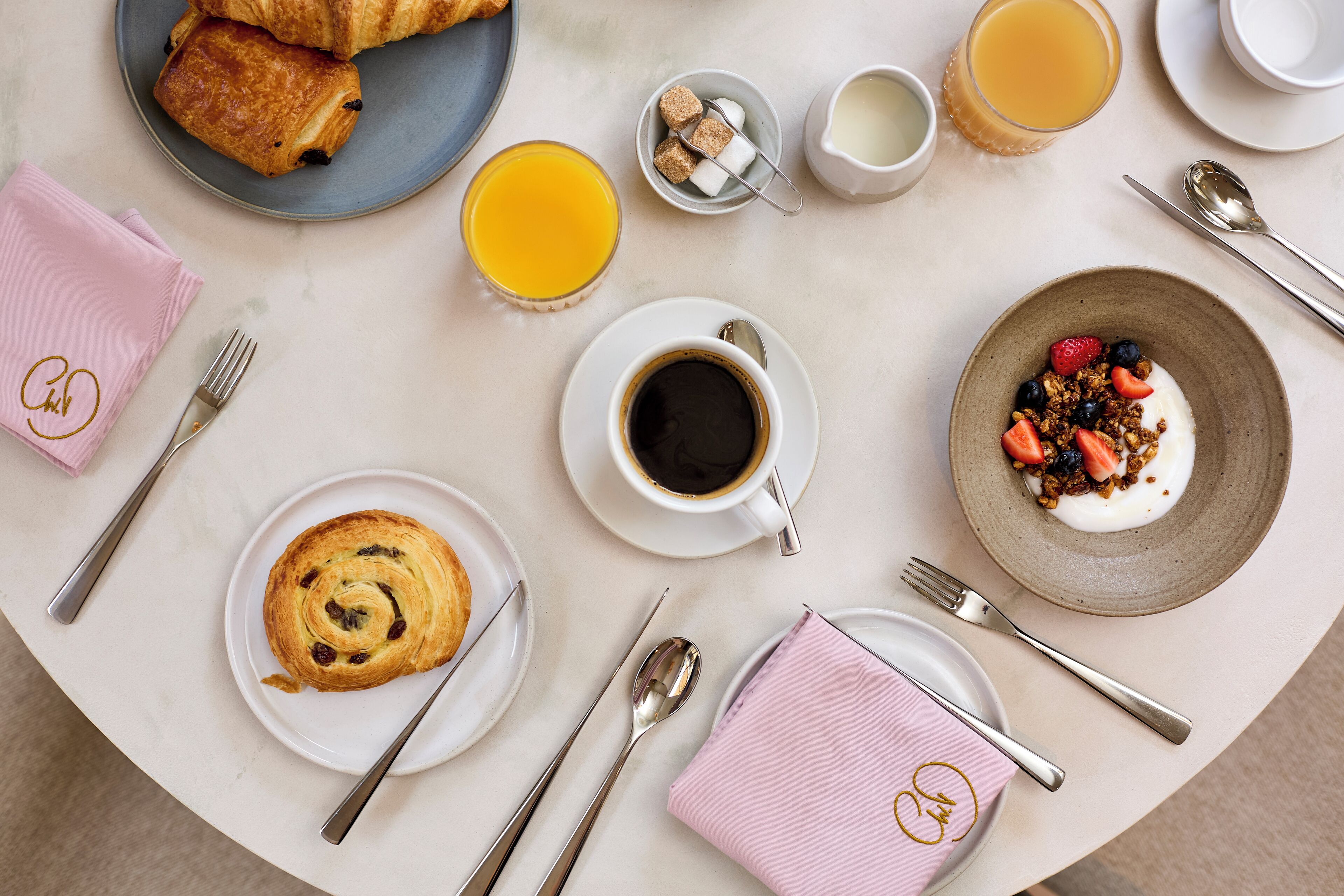 Un desayuno con croissants, un tazón de granola y yogur, un vaso de jugo de naranja y una taza de café sobre una mesa redonda.