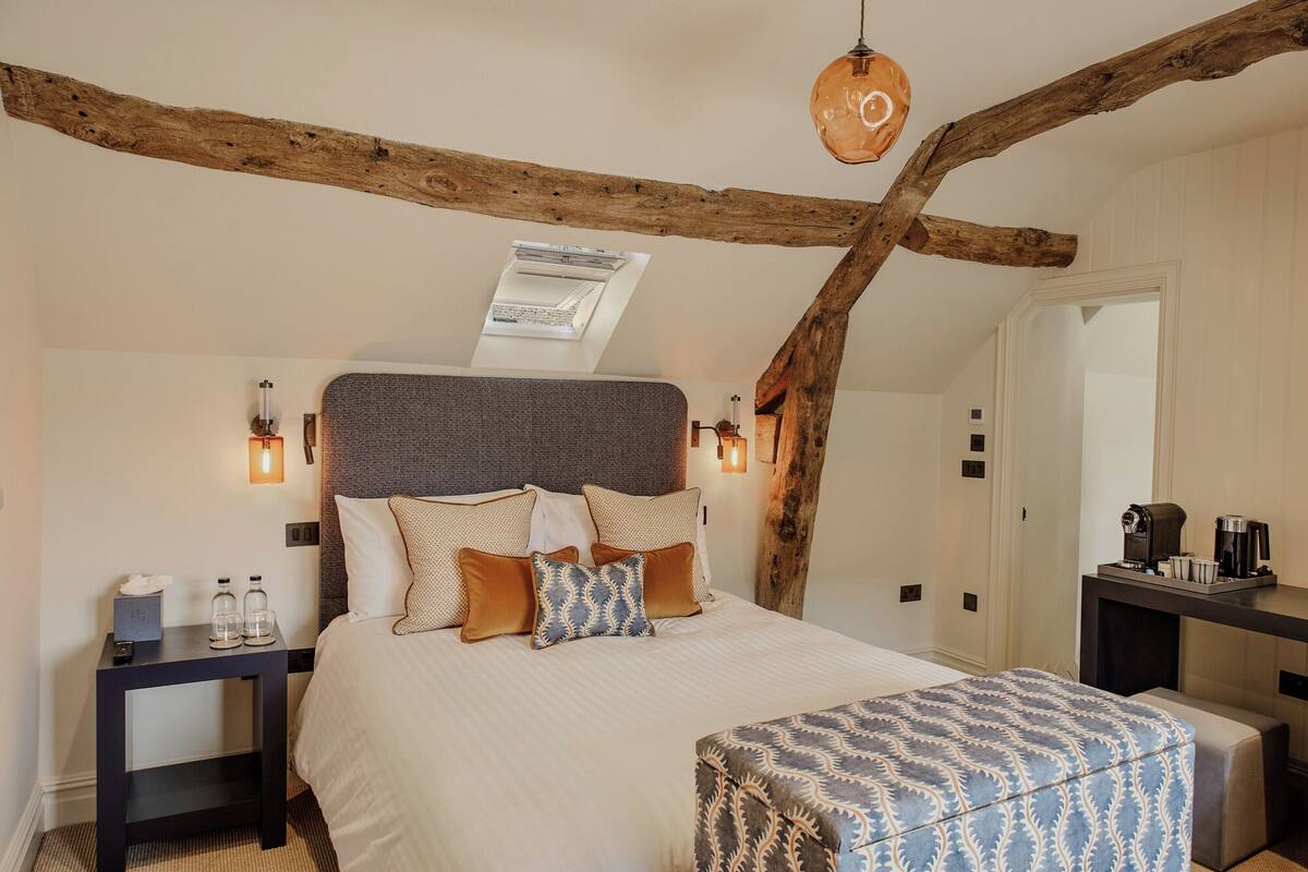 Um quarto com teto inclinado, vigas de madeira aparentes, cama com várias almofadas, mesas de cabeceira e uma pequena mesa com uma cafeteira.
