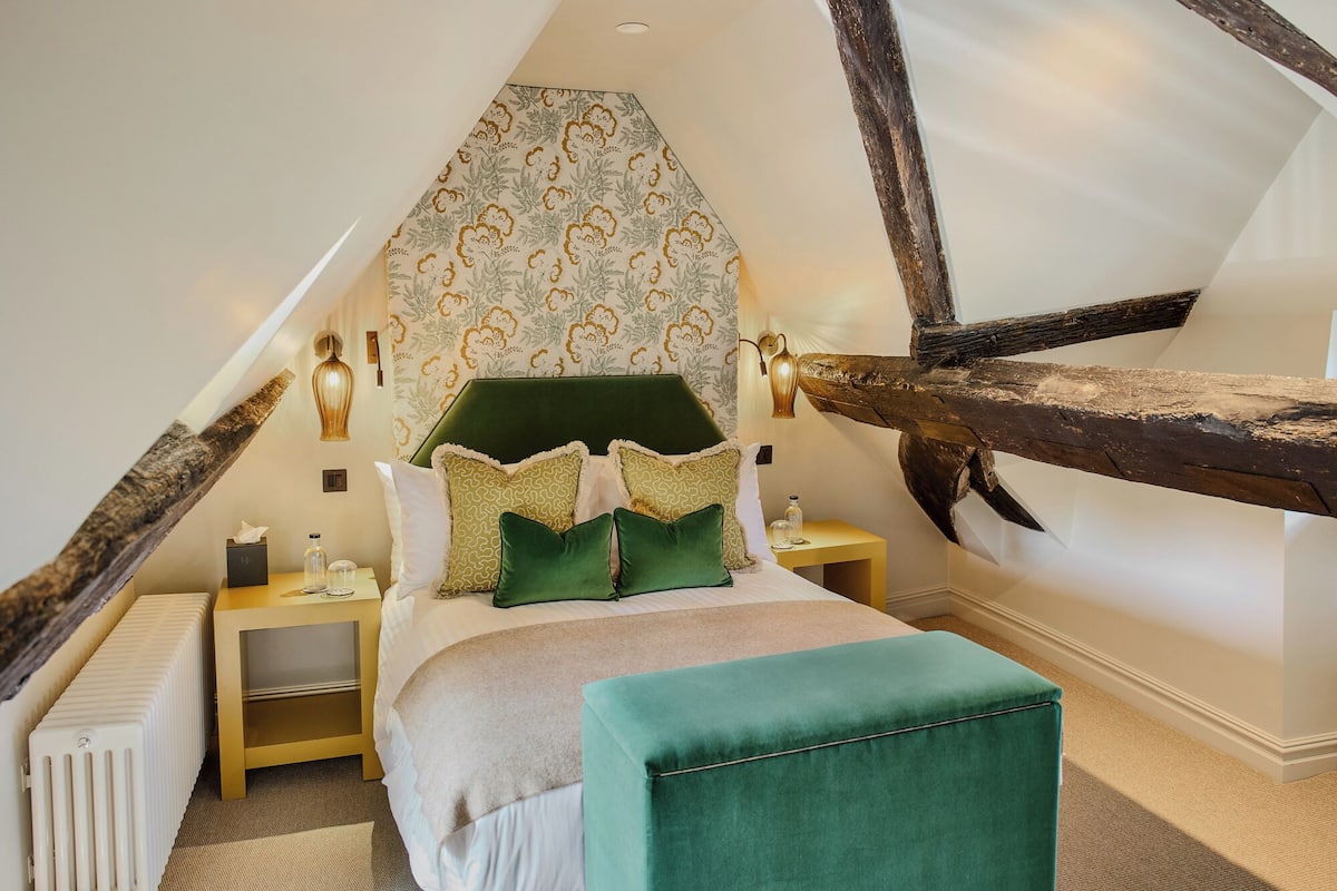 Quarto com teto inclinado, cabeceira estofada em verde, cama com roupa de cama branca, duas mesas de cabeceira com abajures e estrutura de vigas de madeira.