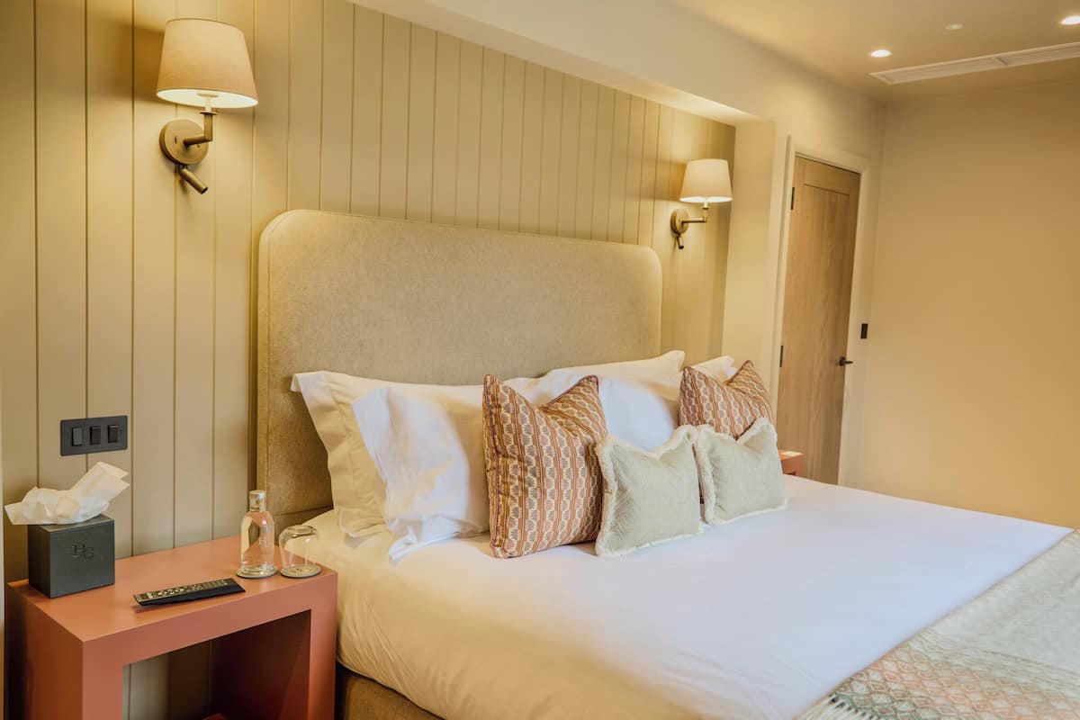 Quarto de hotel com uma cama grande, duas luminárias de cabeceira, mesa de cabeceira com controle remoto, caixa de lenços e um copo de água.