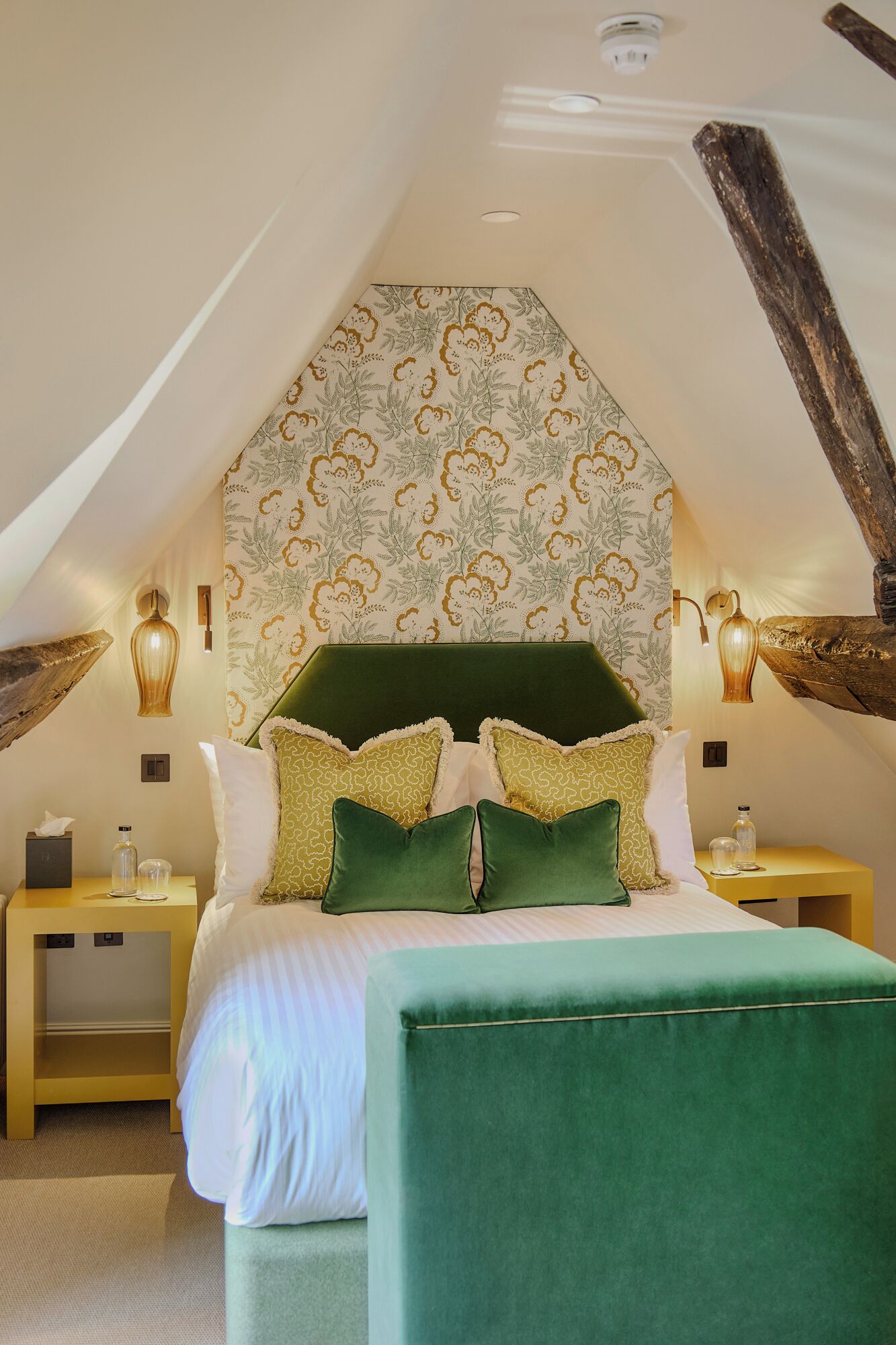 Quarto com cabeceira estofada verde, papel de parede estampado e duas mesas de cabeceira com luminárias.