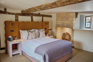 A bedroom with a rustic wooden ceiling, a stone wall, a bed with patterned pillows, a bedside table with a tissue box, and a nightstand with a lamp. - House of George W. Davies (Broadway)