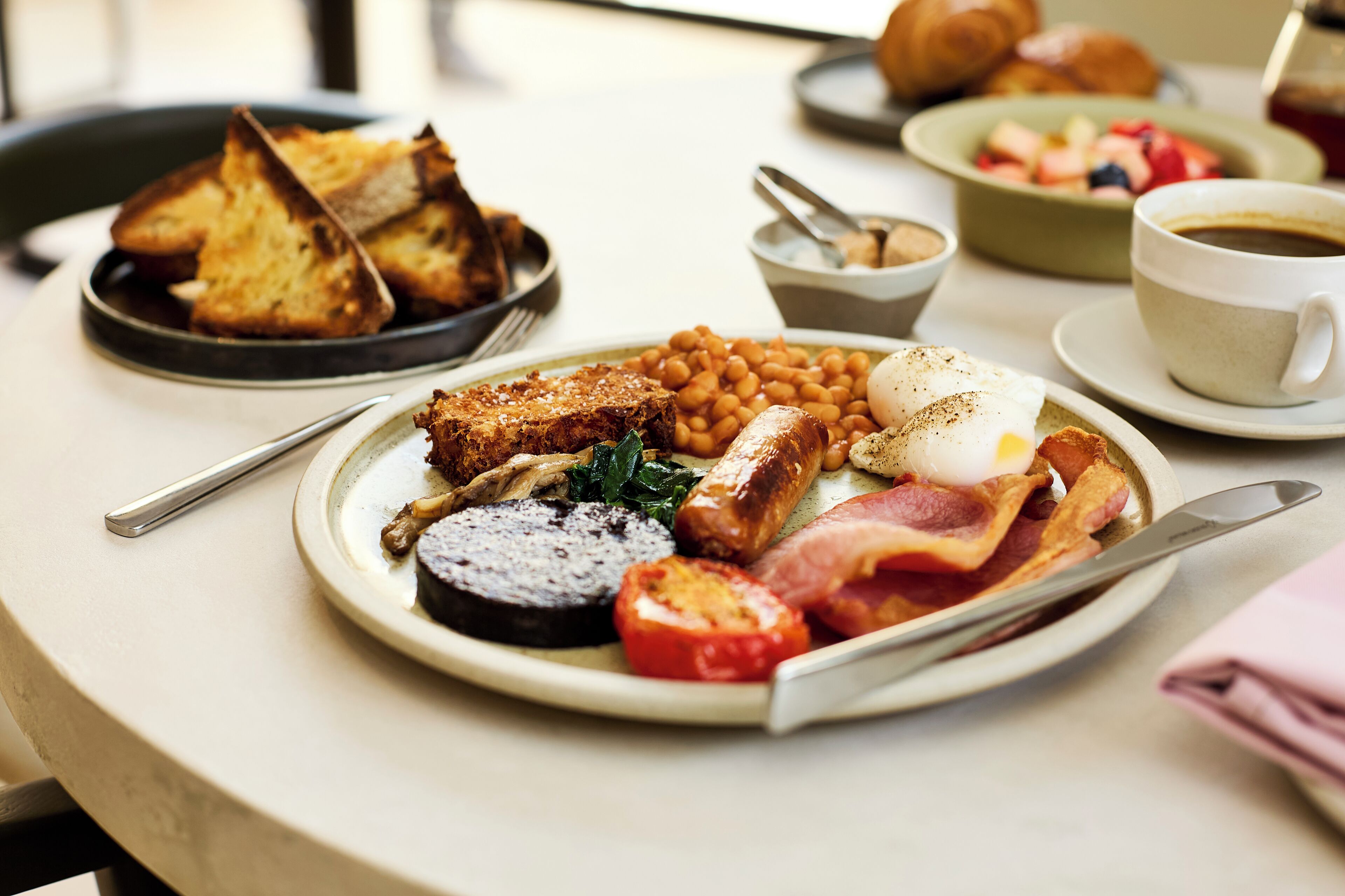 Une table dressée pour le petit-déjeuner avec des toasts, des haricots, des œufs, des tomates et du bacon sur une assiette, ainsi qu’une tasse de café et des croissants en arrière-plan.
