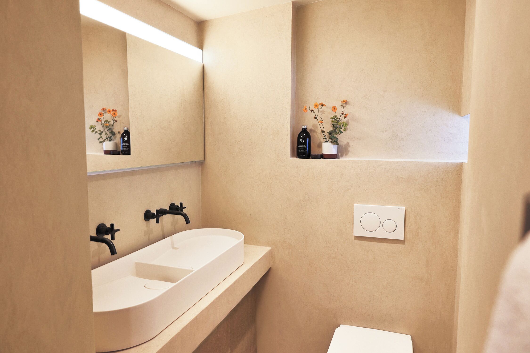 A modern bathroom with a white sink, black faucets, a mirror, and a thermostat on the wall.