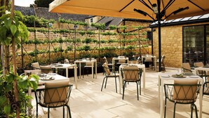 An outdoor patio area with tables and chairs, a large umbrella, and a stone wall with a vineyard in the background. - House of George W. Davies (Broadway)