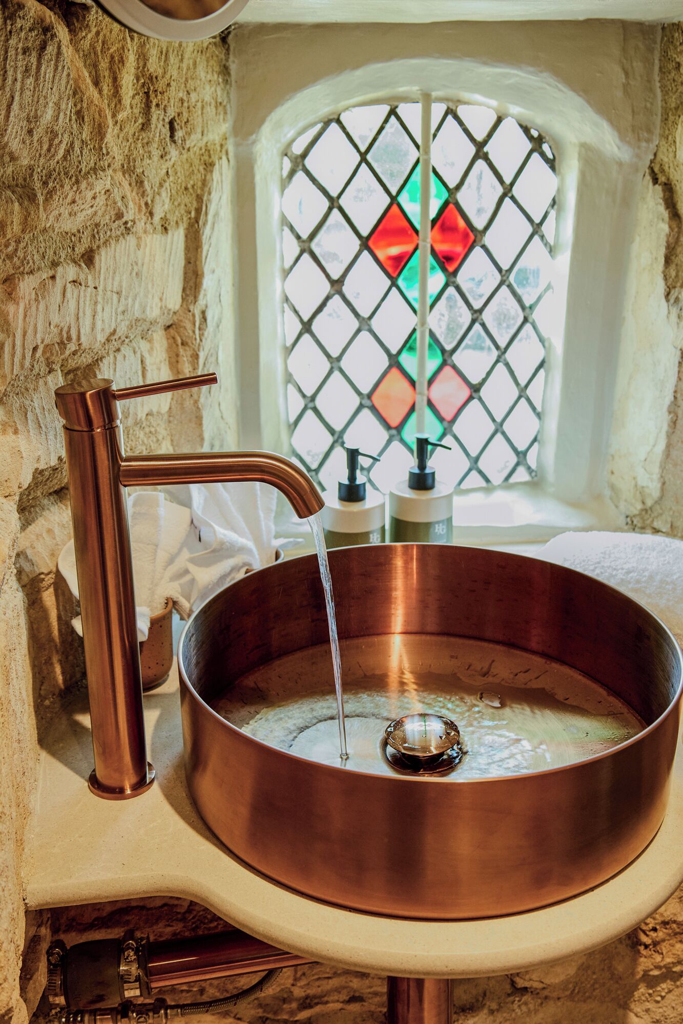 A copper sink with running water, a faucet, and a window with stained glass.