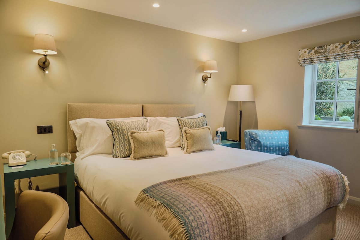 Quarto de hotel com uma cama grande, duas luminárias de cabeceira, mesa de cabeceira com telefone, uma cadeira e janela com vista para área verde.