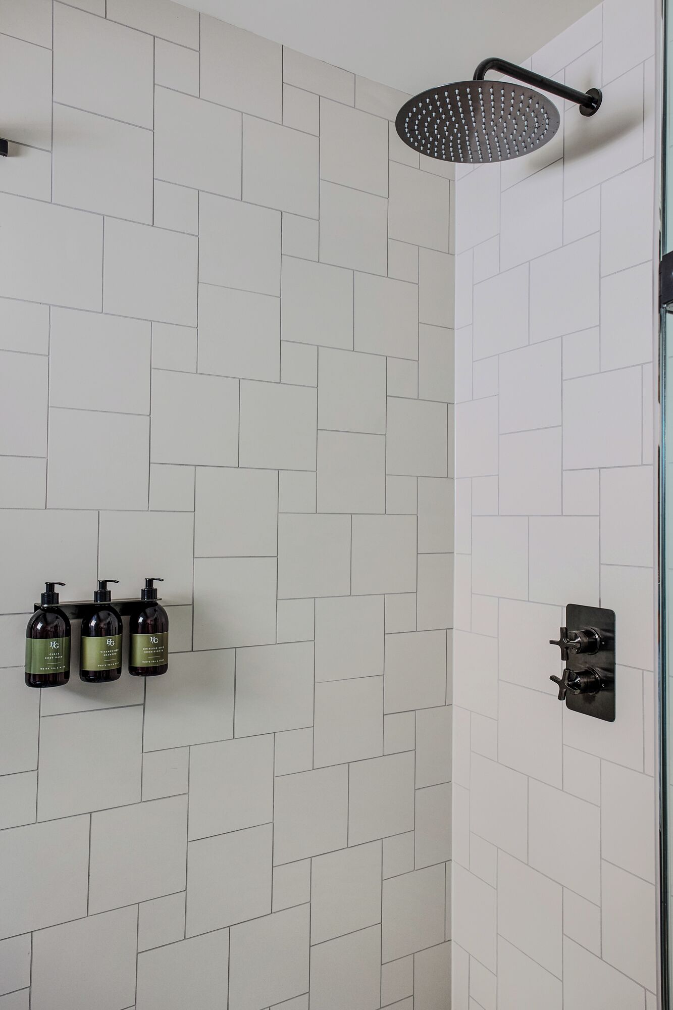 A shower with a black handheld showerhead, white tiled walls, and three bottles of shampoo on a shelf.