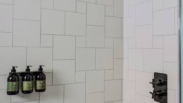 A shower with a black handheld showerhead, white tiled walls, and three bottles of shampoo on a shelf.