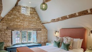 A bedroom with a stone wall, a sloped ceiling, a bed with multiple pillows, a bench, and a small table. - House of George W. Davies (Broadway)