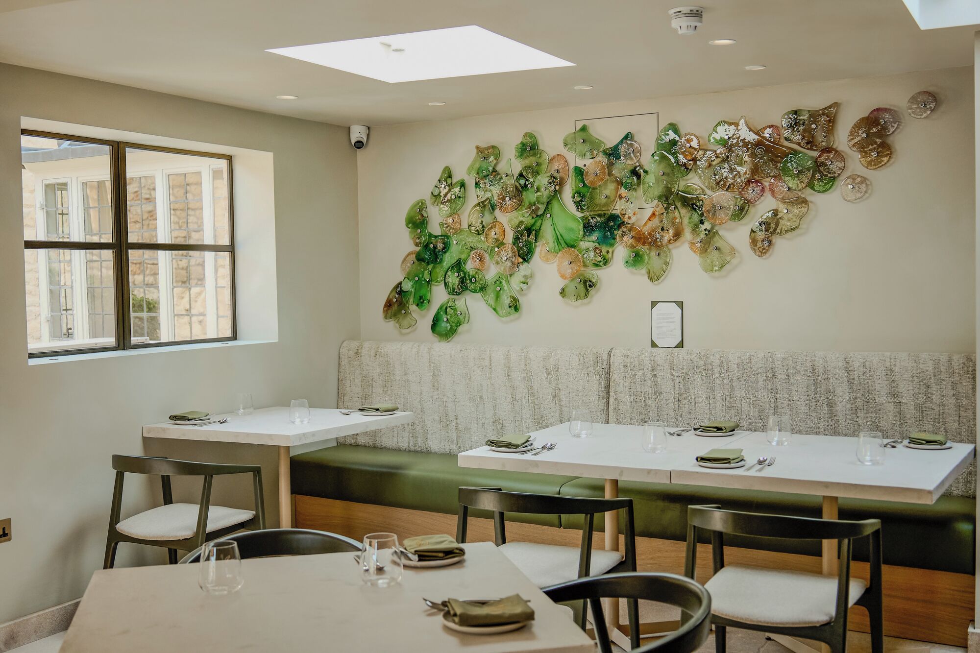 A dining area with a long table, chairs, and a bench. There is a window with multiple panes and a wall-mounted security camera.