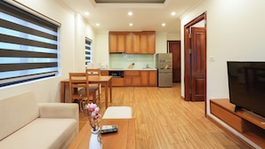 A modern kitchen with wooden cabinets, a dining area with a table and chairs, a sofa, and a TV on a wooden stand. - NINE HOUSING 68 Kim Ma Thuong (Hanoi)