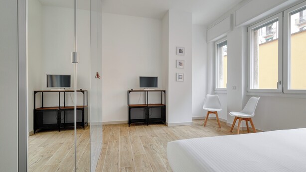 Un dormitorio moderno con una cama, dos escritorios con computadoras, una silla y ventanas amplias.