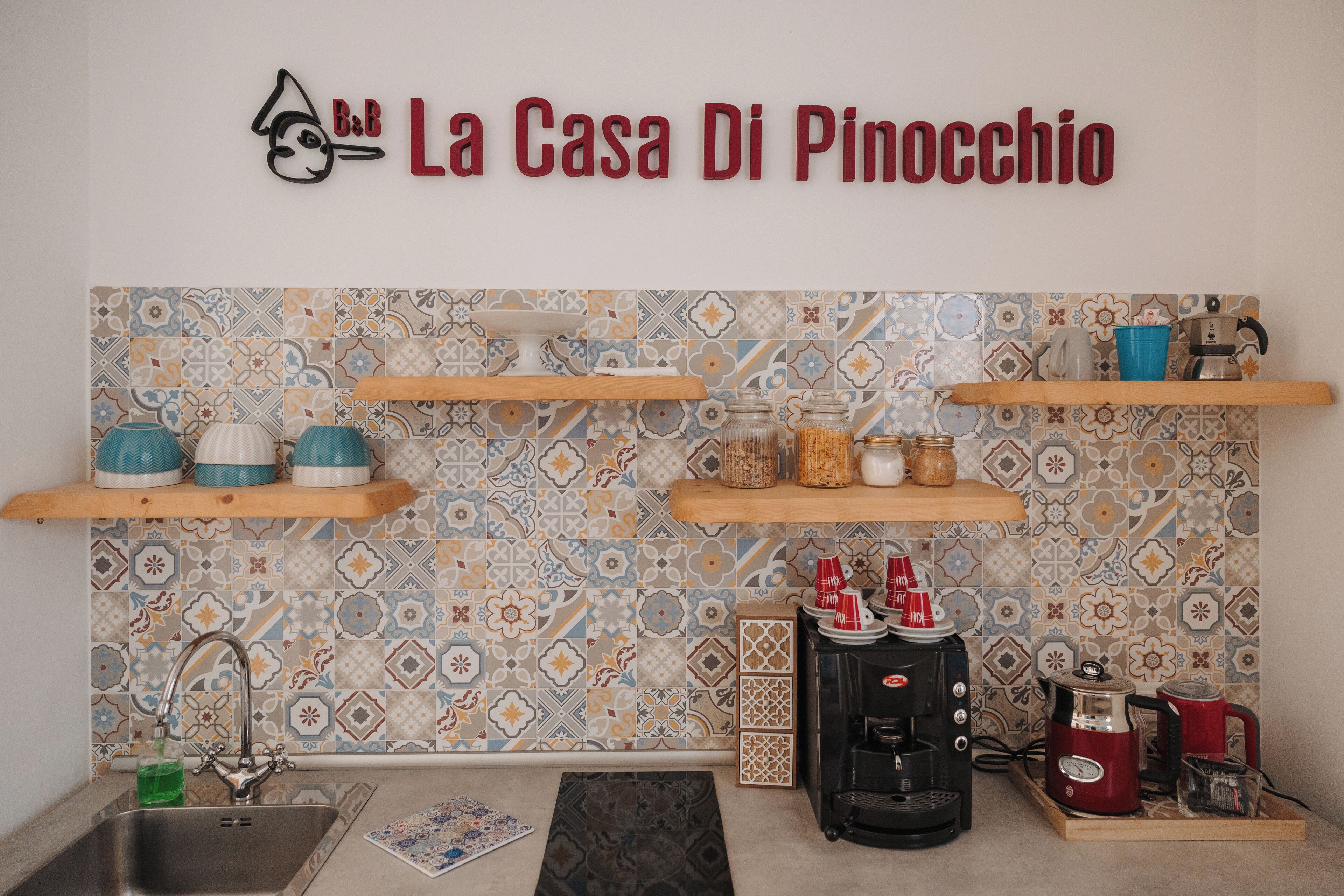 Una cocina con salpicadero de azulejos, estantes de madera, una cafetera y un lavamanos.
