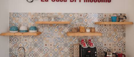 Una cocina con salpicadero de azulejos, estantes de madera, una cafetera y un lavamanos.