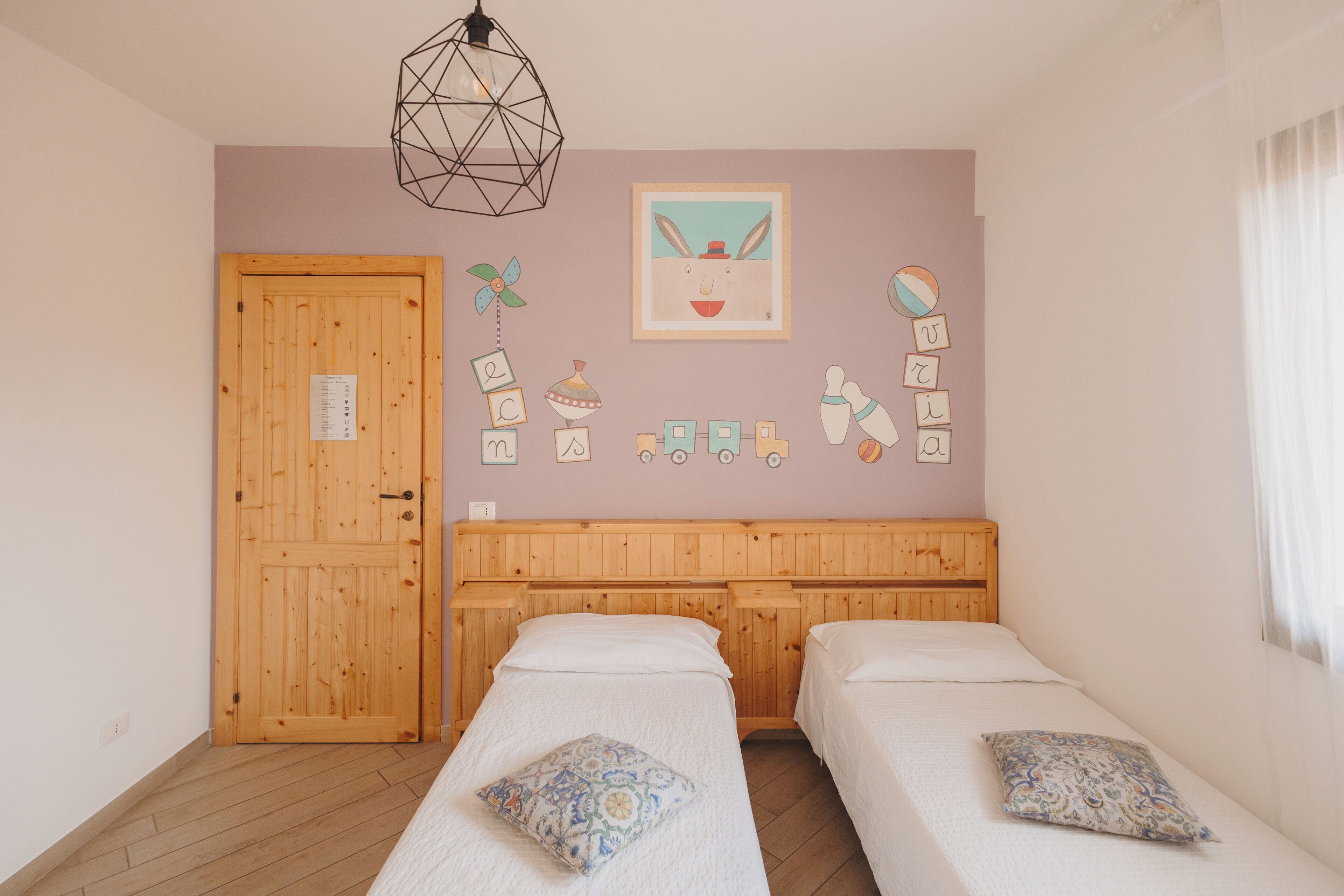 Une chambre avec deux lits simples, des têtes de lit en bois, un luminaire suspendu aux motifs géométriques, un mur décoré de lettres de l’alphabet et une porte sur laquelle est affiché un avis.