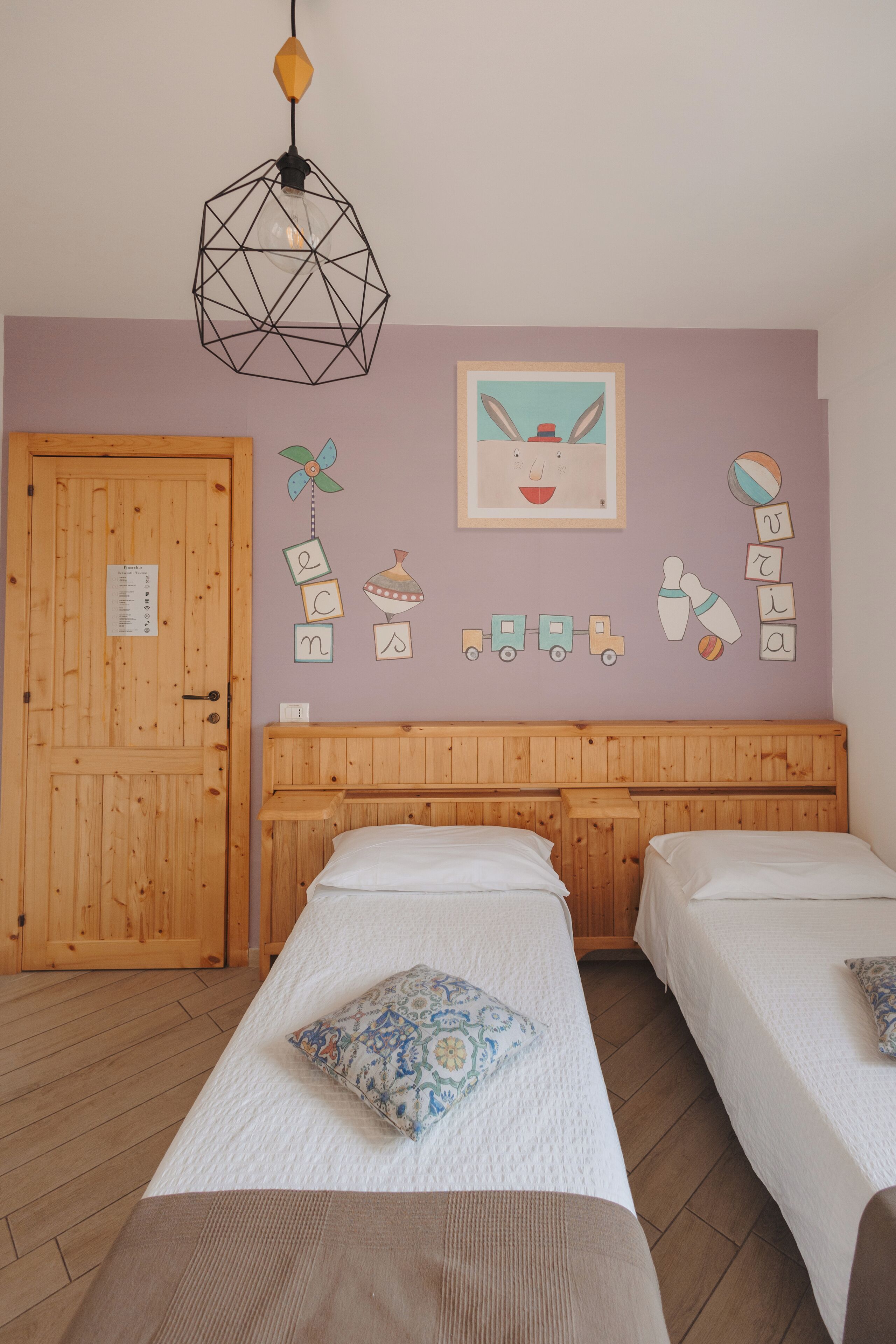 Ein Schlafzimmer mit zwei Betten, einem Holzkopfstück, einer geometrischen Pendelleuchte und einer Wanddekoration mit bunten Kinderbuchstaben und einem Bilderrahmen.