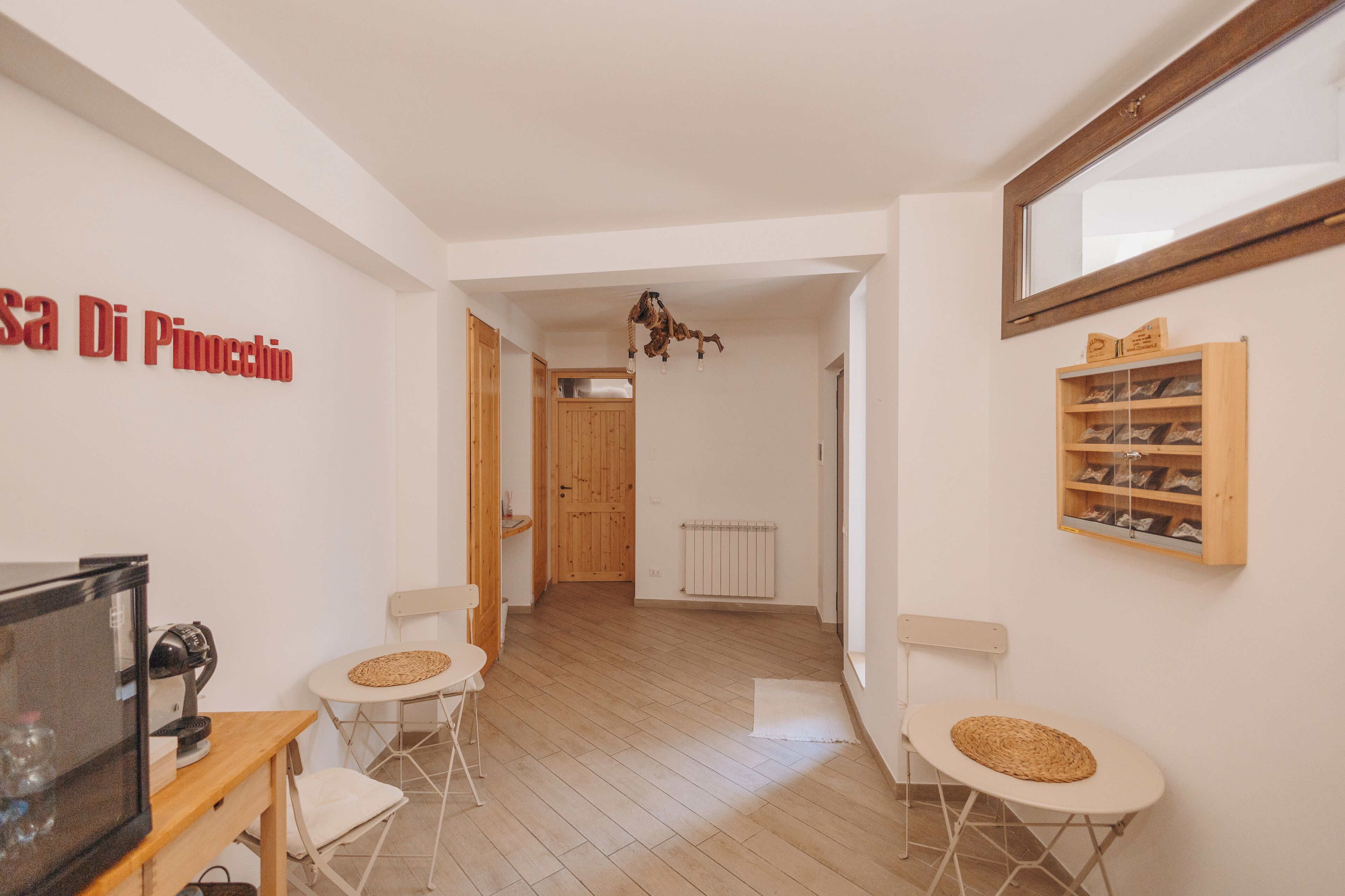 Una habitación con piso de madera, paredes blancas, una puerta de madera, una mesita con una silla, un estante con zapatos y un letrero que dice "Casa Di Pinocchio".