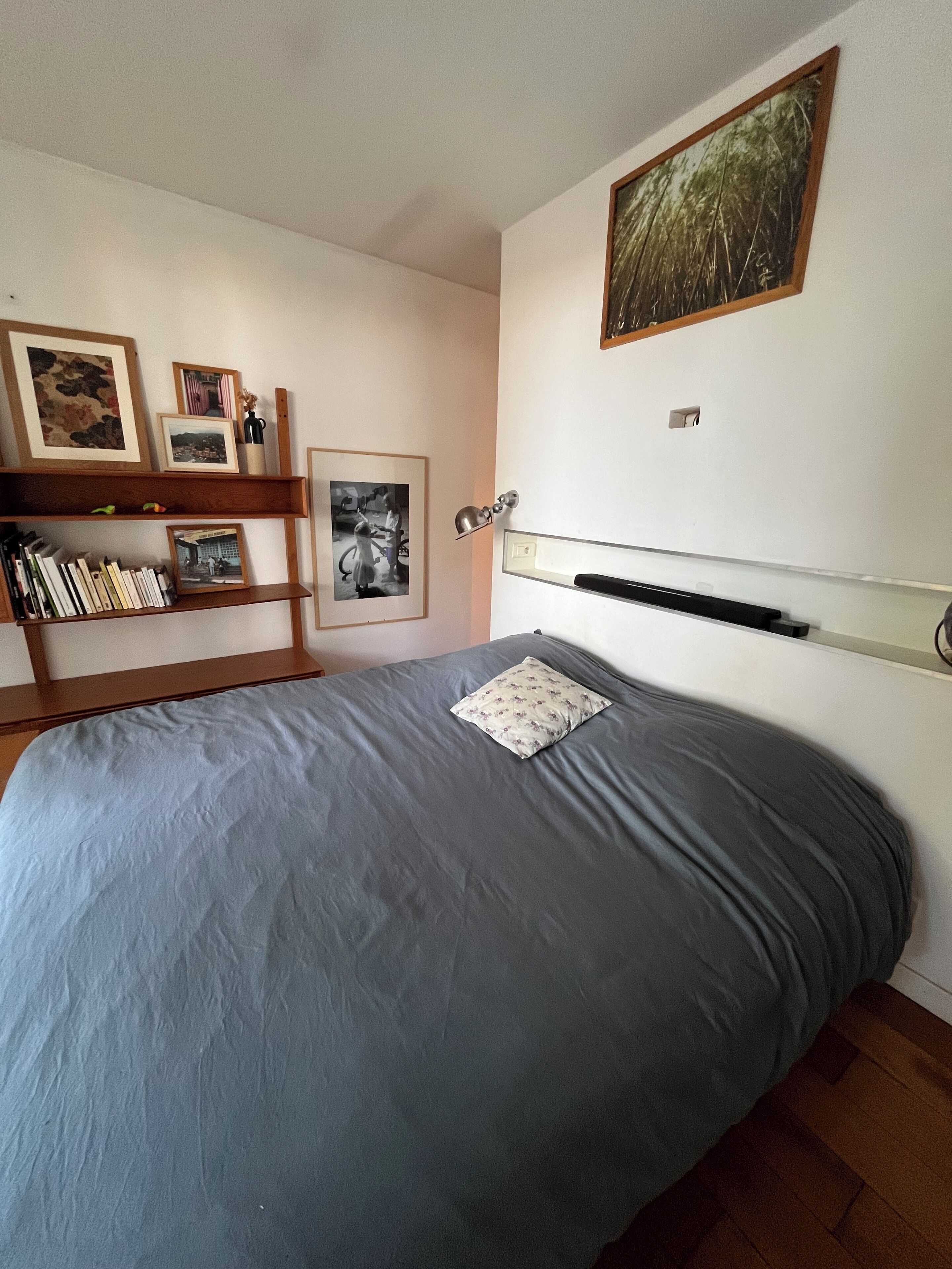 A bedroom with a bed, a wooden shelf with books and pictures, and a wall-mounted picture frame.