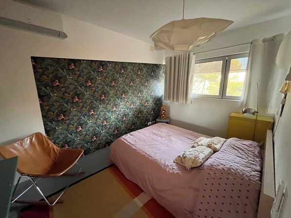 Una camera da letto con una testiera a motivi decorativi, un letto con biancheria rosa, una sedia, un comodino con una lampada e una finestra con tende.