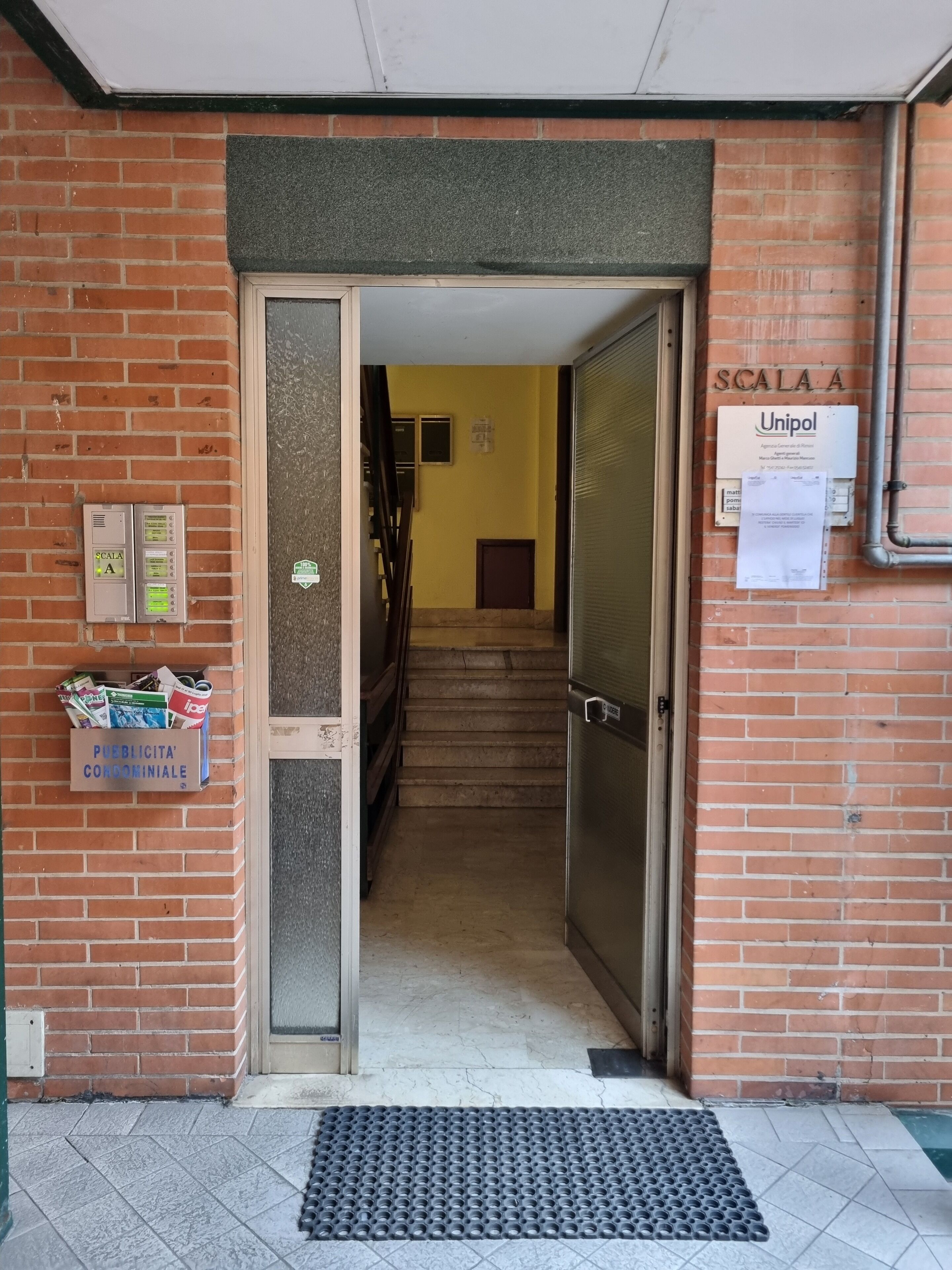 Ein Gebäudeeingang mit Backsteinfassade, einer Metalttür, einer Treppe, die nach innen führt, und einem Schild mit 'Unipol' auf der rechten Seite.